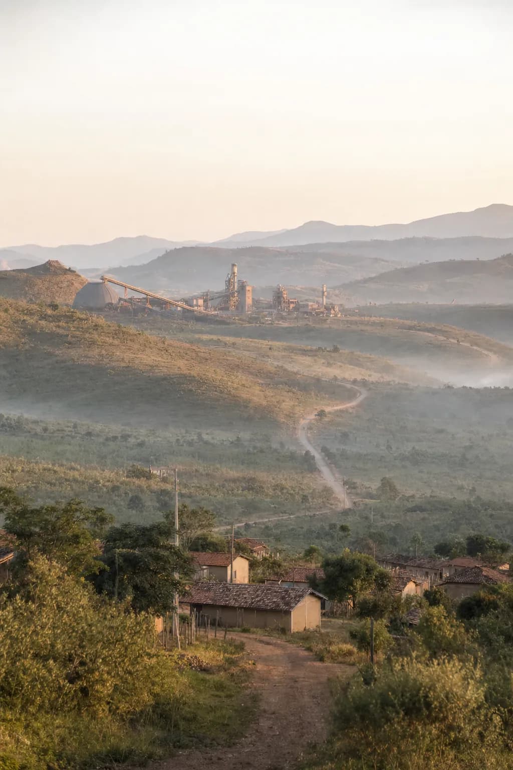 Vale com pequena comunidade rural em primeiro plano e estruturas de mineração distantes no horizonte, sob luz de fim de tarde